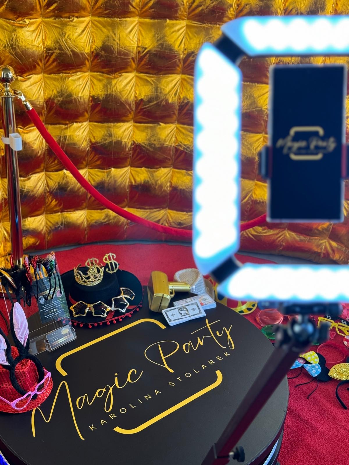 Przygotowana fotobudka z rekwizytami: kapelusz z koroną i symbolami dolara, zabawkowy pistolet, sztuczne pieniądze, okulary, królicze uszy, na tle złotej ściany i czerwonego dywanu.