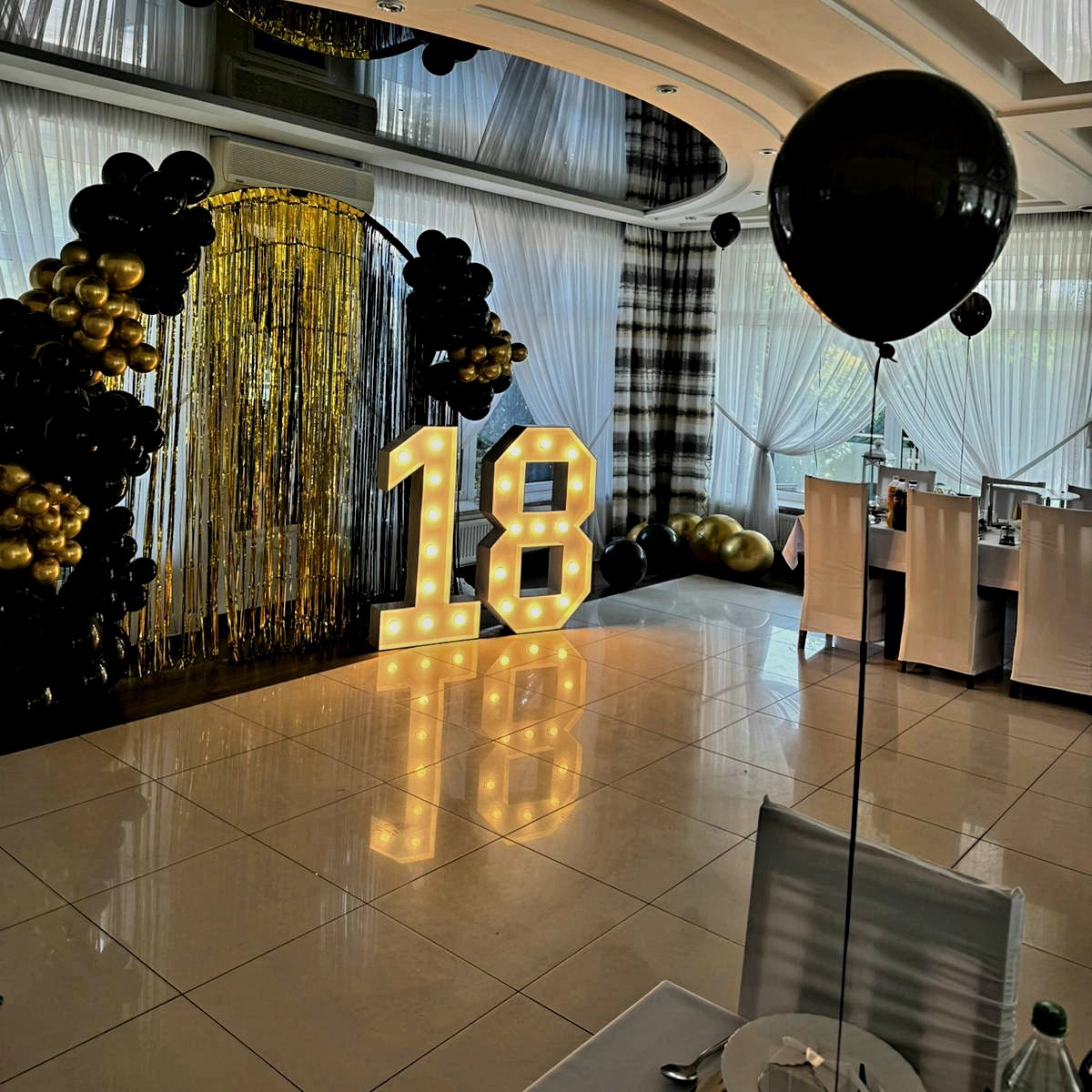 Dekoracja sali na 18 urodziny: podświetlane cyfry '18', złote frędzle, girlanda z czarnych i złotych balonów oraz duży czarny balon na sali z nakrytymi stołami.
