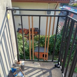 Przedsiębiorstwo Techniczne MTC Wiktor Suska - Czarna balustrada balkonowa z drewnianymi elementami dekoracyjnymi, widok z góry na betonowy balkon z puszką farby, pędzlem i taśmą malarską w słoneczny dzień.