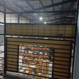 Przedsiębiorstwo Techniczne MTC Wiktor Suska - Metalowa konstrukcja balustrady z widokiem na magazyn z regałami pełnymi kartonów, oświetlona górnym światłem.