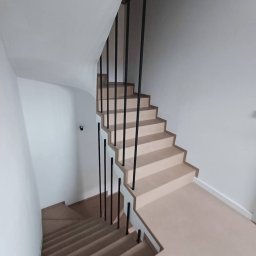 Przedsiębiorstwo Techniczne MTC Wiktor Suska - Nowoczesne schody wewnętrzne z beżowymi stopniami i minimalistyczną, czarną balustradą z pionowych prętów, widok z góry na dół biegu schodów.