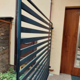 Nowoczesna, pozioma, metalowa konstrukcja ogrodzeniowa w kolorze grafitowym, zamontowana przed budynkiem z beżową elewacją i brązowymi drzwiami.