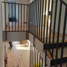 Nowoczesna, czarna poręcz schodowa z pionowymi prętami, widok z góry, na schodach widoczne zabezpieczenia podczas prac wykończeniowych, w tle widoczne lampy budowlane i materiały.