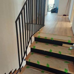 Przedsiębiorstwo Techniczne MTC Wiktor Suska - Nowoczesne schody z czarną metalową balustradą, zabezpieczone kartonem i taśmą malarską na czas remontu, widok z dołu.