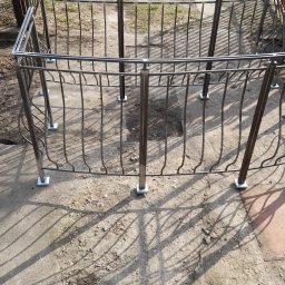 Przedsiębiorstwo Techniczne MTC Wiktor Suska - Metalowa balustrada balkonowa z pionowymi prętami o zakrzywionym kształcie, zamontowana na betonowej powierzchni, rzucająca wyraźny cień w słoneczny dzień.