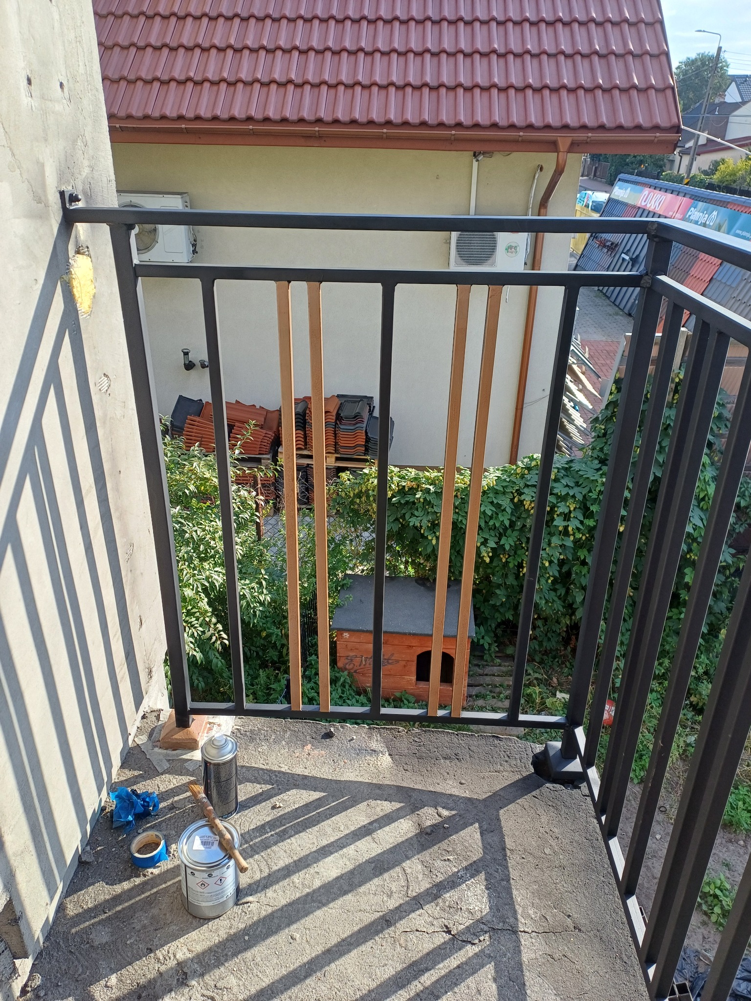 Czarna balustrada balkonowa z drewnianymi elementami dekoracyjnymi, widok z góry na betonowy balkon z puszką farby, pędzlem i taśmą malarską w słoneczny dzień.