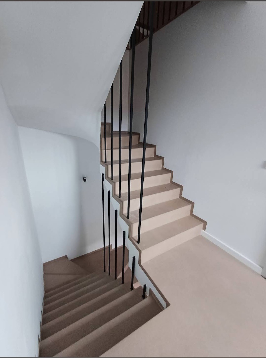 Nowoczesne schody wewnętrzne z beżowymi stopniami i minimalistyczną, czarną balustradą z pionowych prętów, widok z góry na dół biegu schodów.