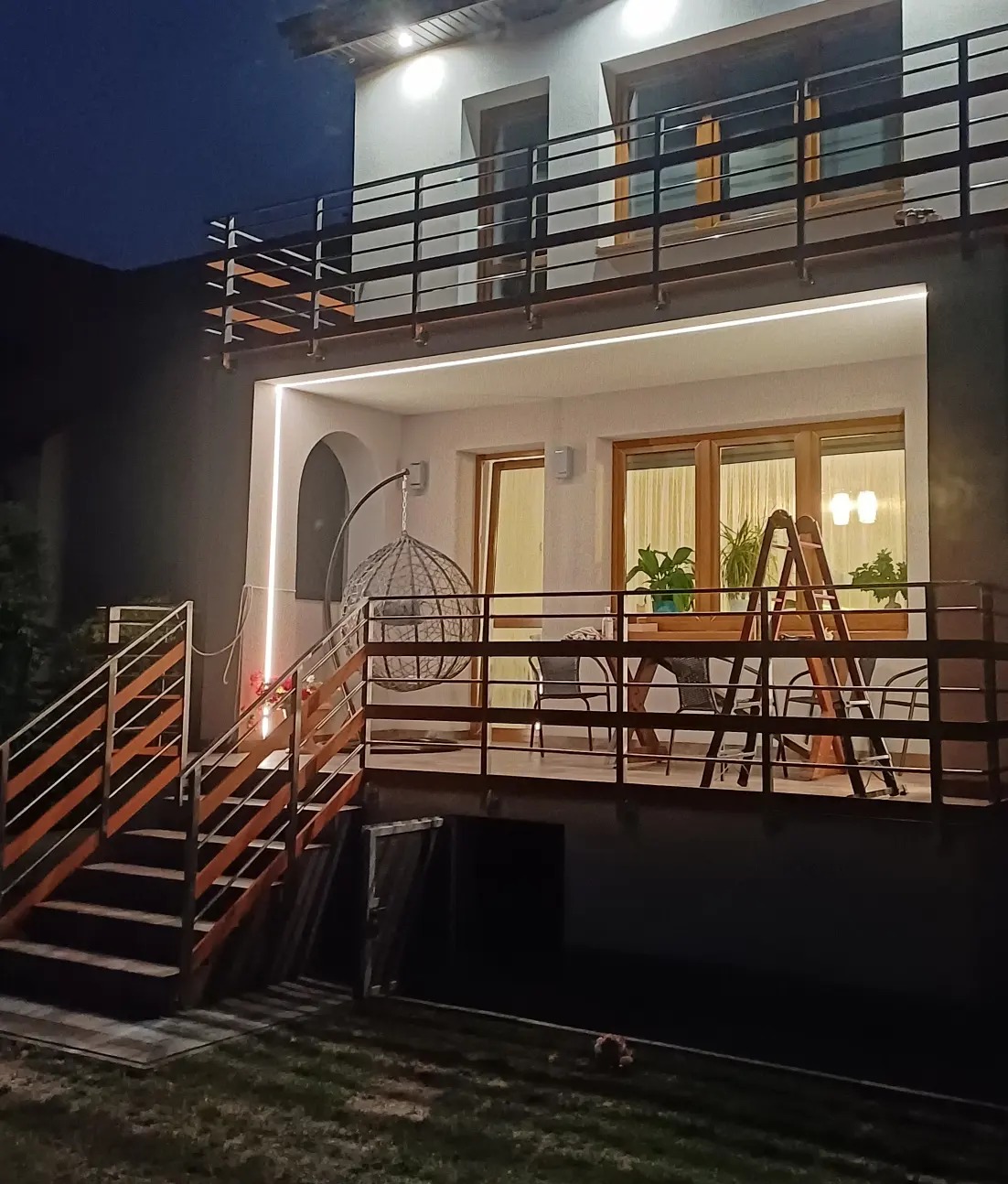 Dwupoziomowy taras z drewnianymi schodami i balustradami, oświetlony taśmą LED, z wiszącym fotelem i widokiem na dom nocą.