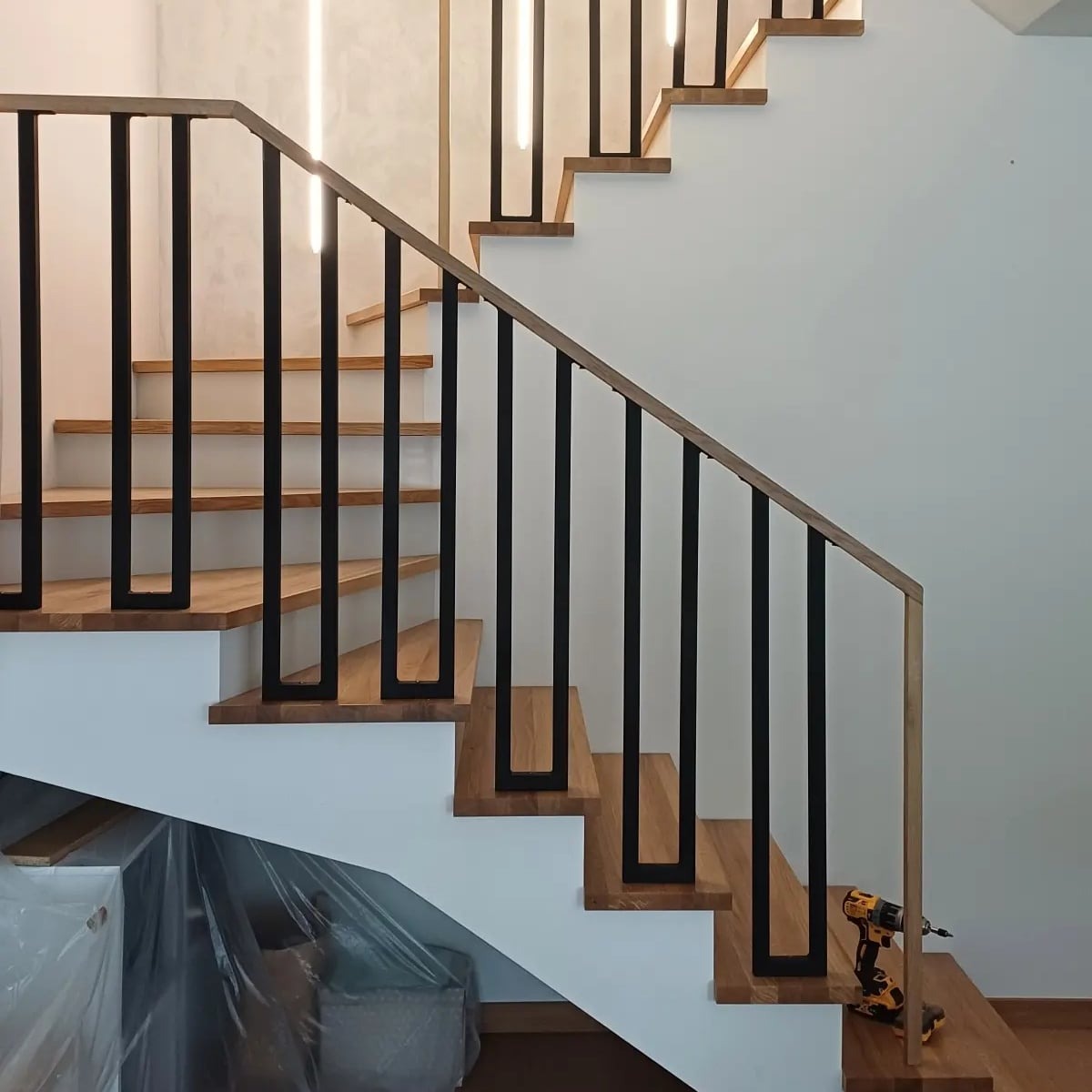 Drewniane schody z czarną, metalową balustradą o minimalistycznym designie, widoczna wkrętarka akumulatorowa na jednym ze stopni.