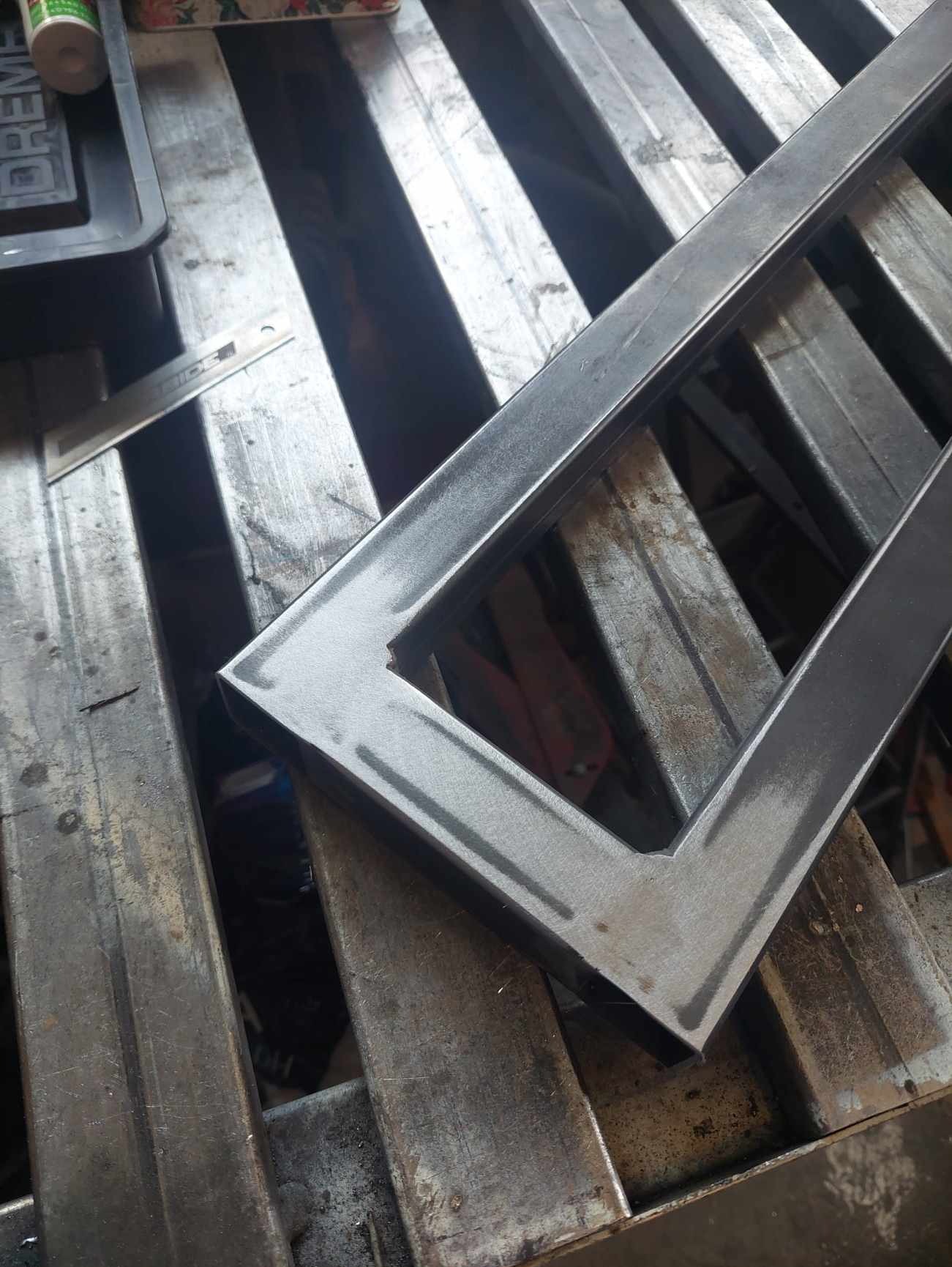 Metalowa rama o prostokątnym kształcie z wyciętym narożnikiem, leżąca na warsztatowym stole z profili, widoczne ślady obróbki i niedoskonałości powierzchni.
