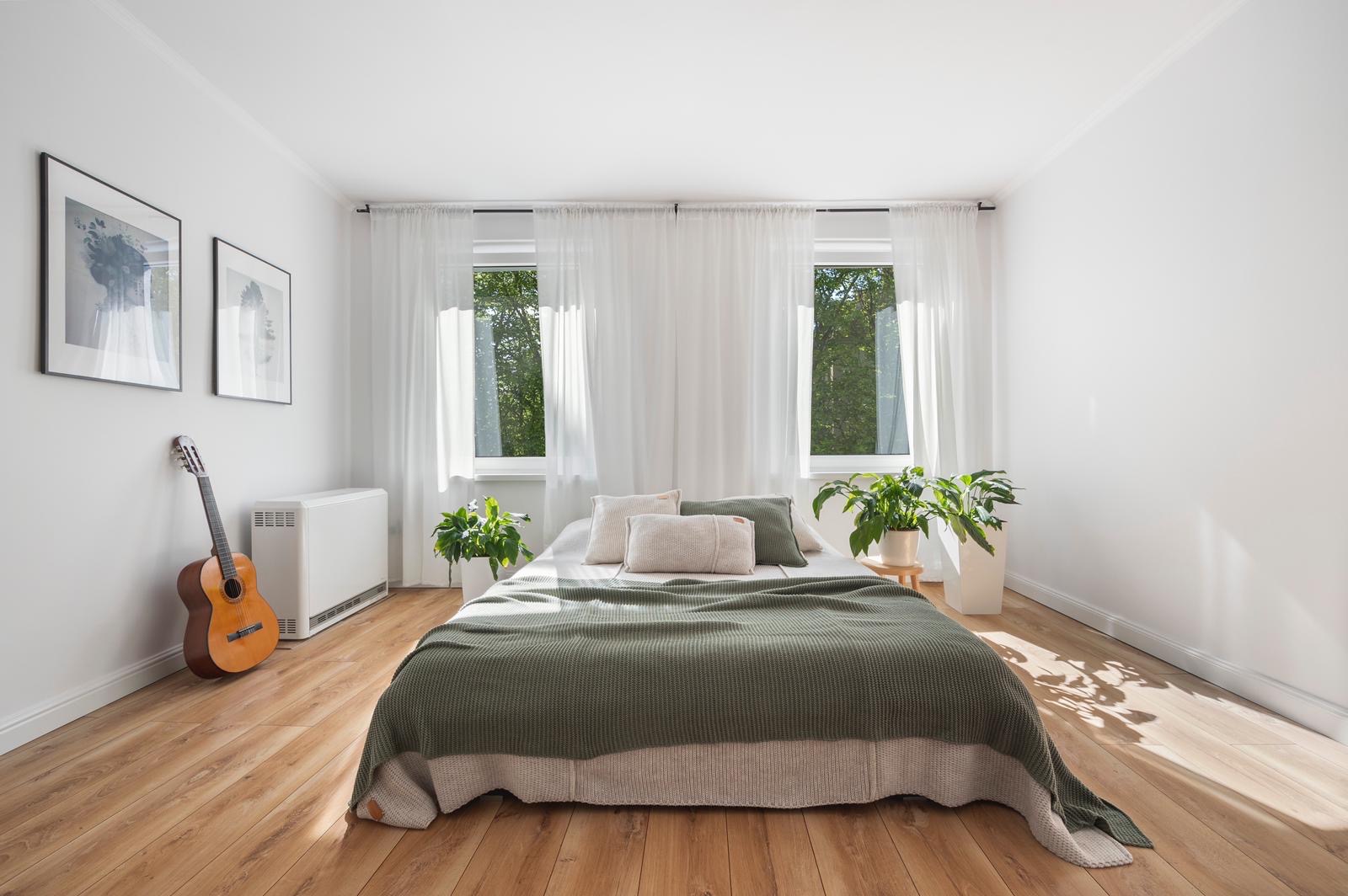 Jasne wnętrze sypialni z drewnianą podłogą, łóżkiem z zieloną narzutą i białymi poduszkami, dwoma oknami z białymi zasłonami, gitarą akustyczną opartą o ścianę obok grzejnika oraz roślinami...