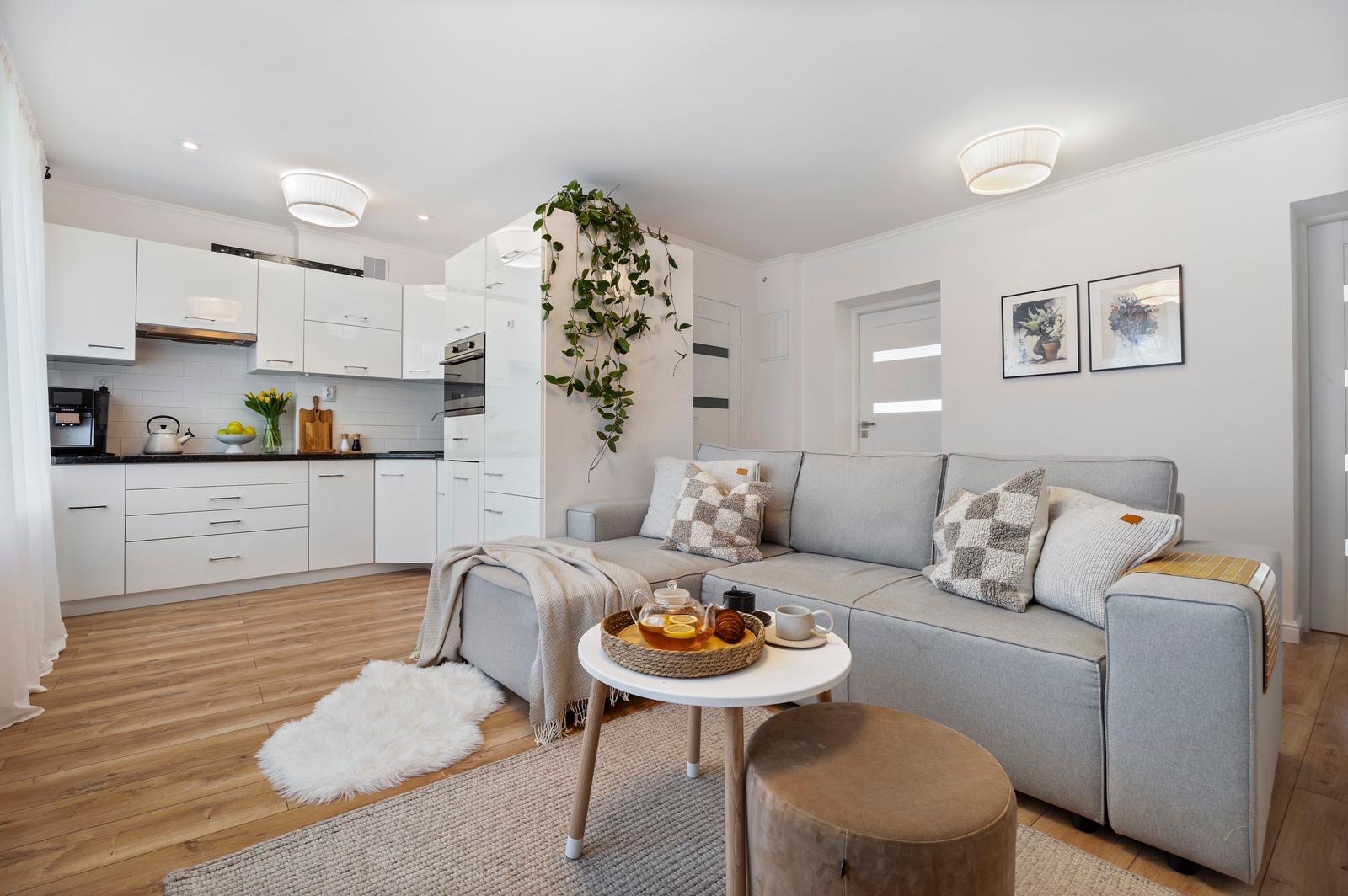 Jasne wnętrze mieszkania z szarą sofą narożną, białymi meblami kuchennymi i drewnianą podłogą, dekorowane roślinami i dodatkami w neutralnych kolorach.