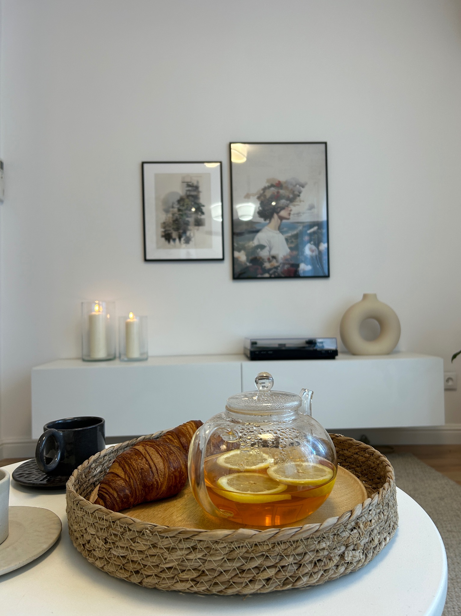 Stół z wiklinową tacą, na której znajduje się szklany czajnik z herbatą cytrynową i rogalik, w tle minimalistyczny salon z białymi ścianami, grafikami w czarnych ramkach i białym kredensem...