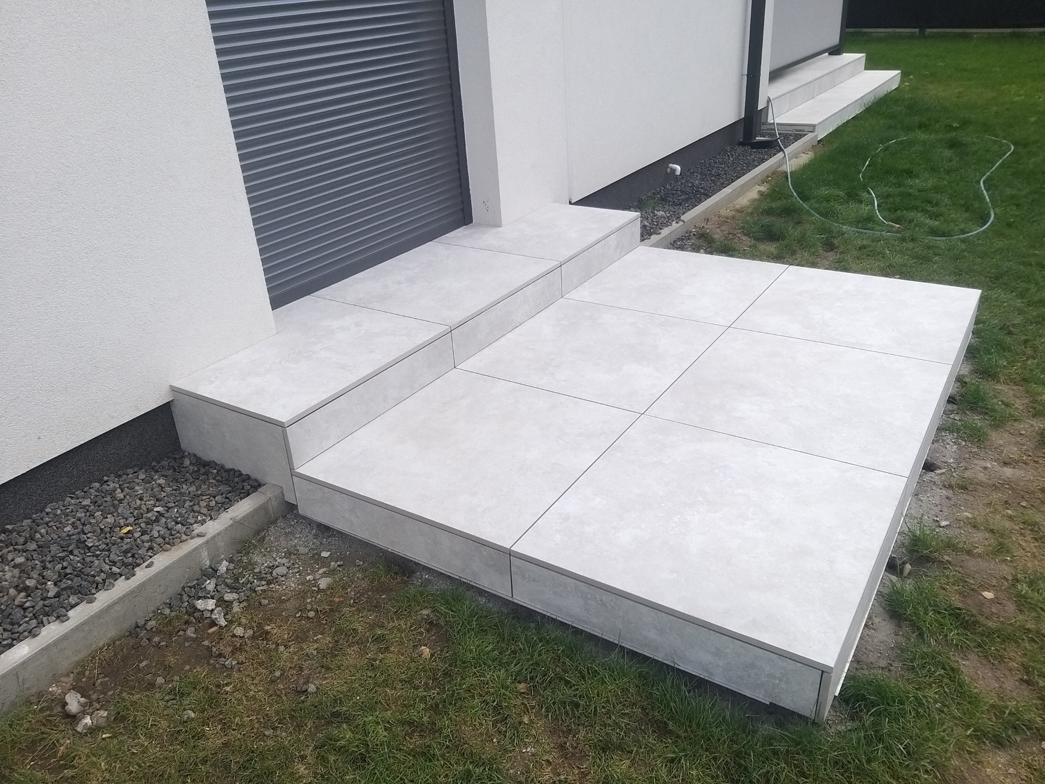 Jasnoszara, kwadratowa płyta tarasowa, ułożona na schodach i podwyższeniu przy budynku z szarą roletą zewnętrzną.