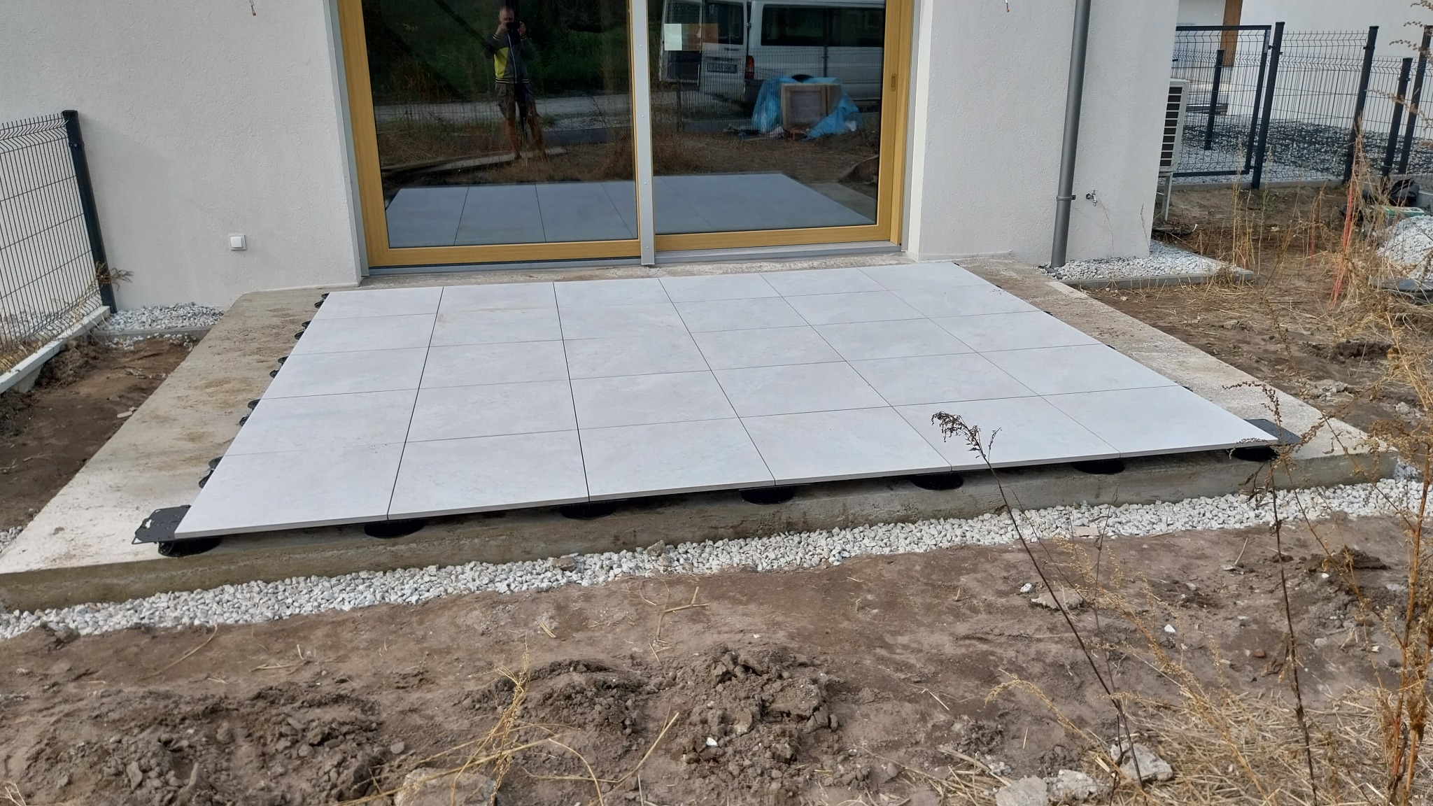Montaż tarasu z jasnych płyt kamiennych na regulowanych wspornikach, widoczny betonowy podkład i drobny żwir wokół, w tle okno przesuwne i odbicie osoby.