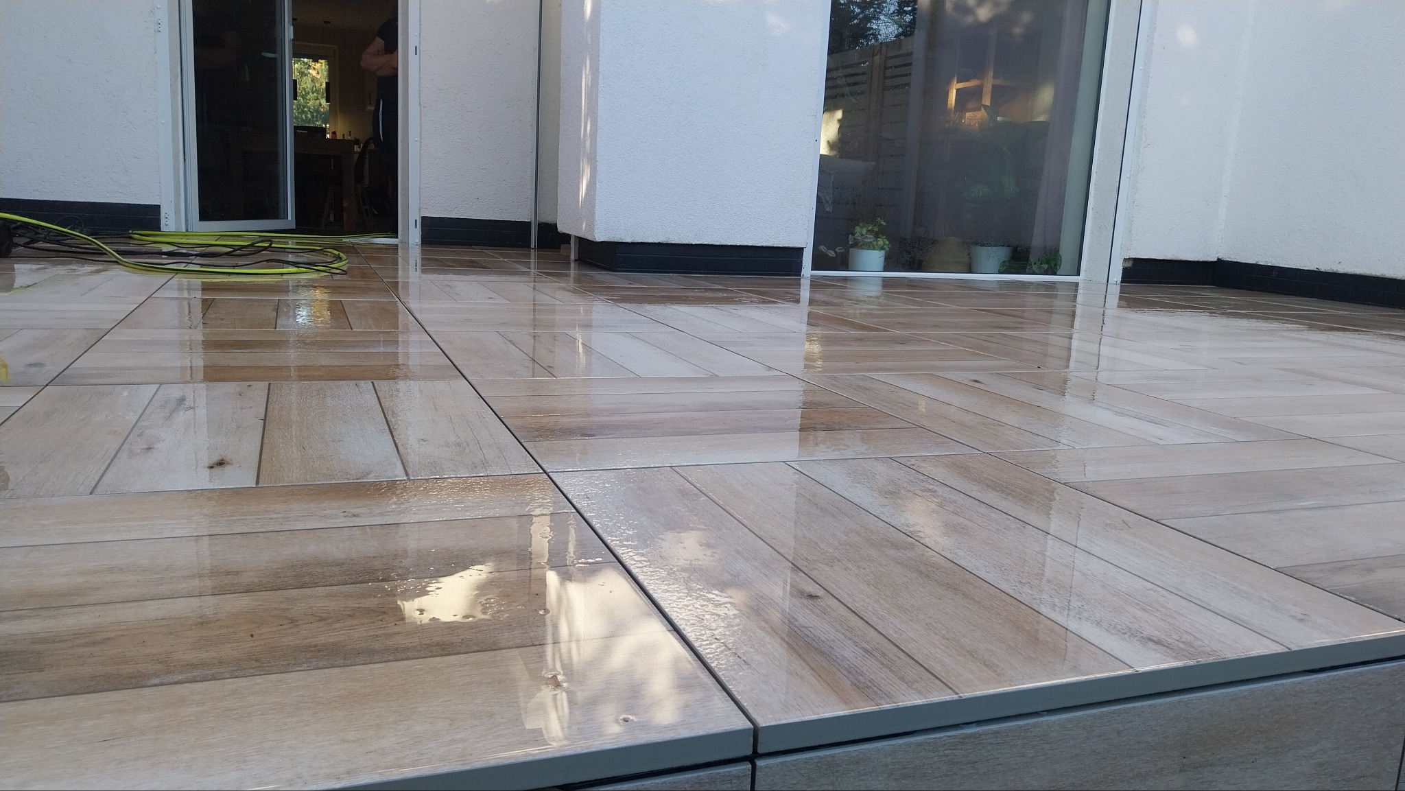 Świeżo umyty taras z płytek imitujących drewno, z widocznymi refleksami światła i fragmentem zwiniętego węża ogrodowego.