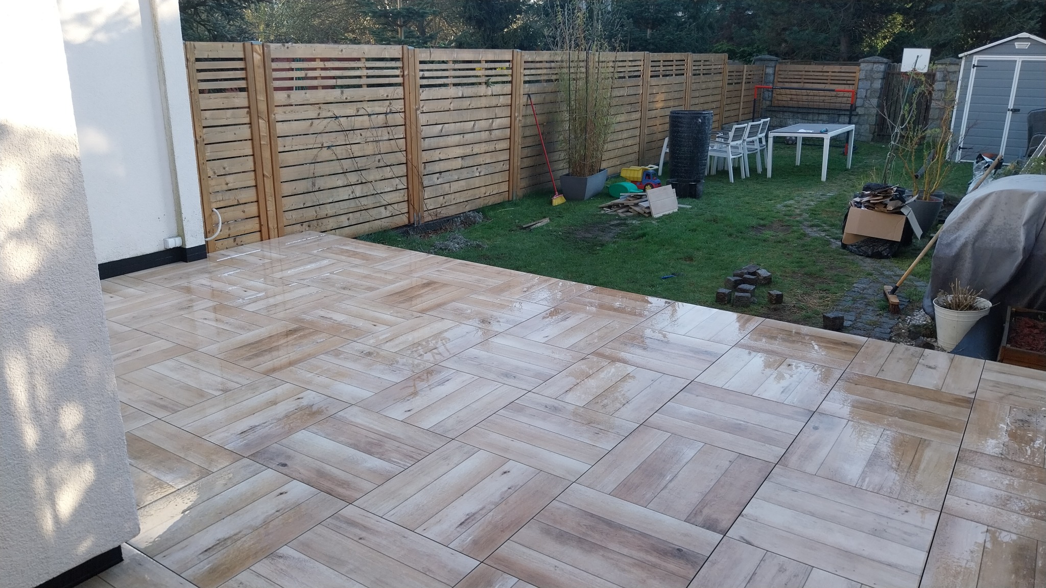 Ułożona z jasnych płytek tarasowych w jodełkę, częściowo mokra powierzchnia tarasu, w tle drewniany płot i trawnik z elementami wyposażenia ogrodu.