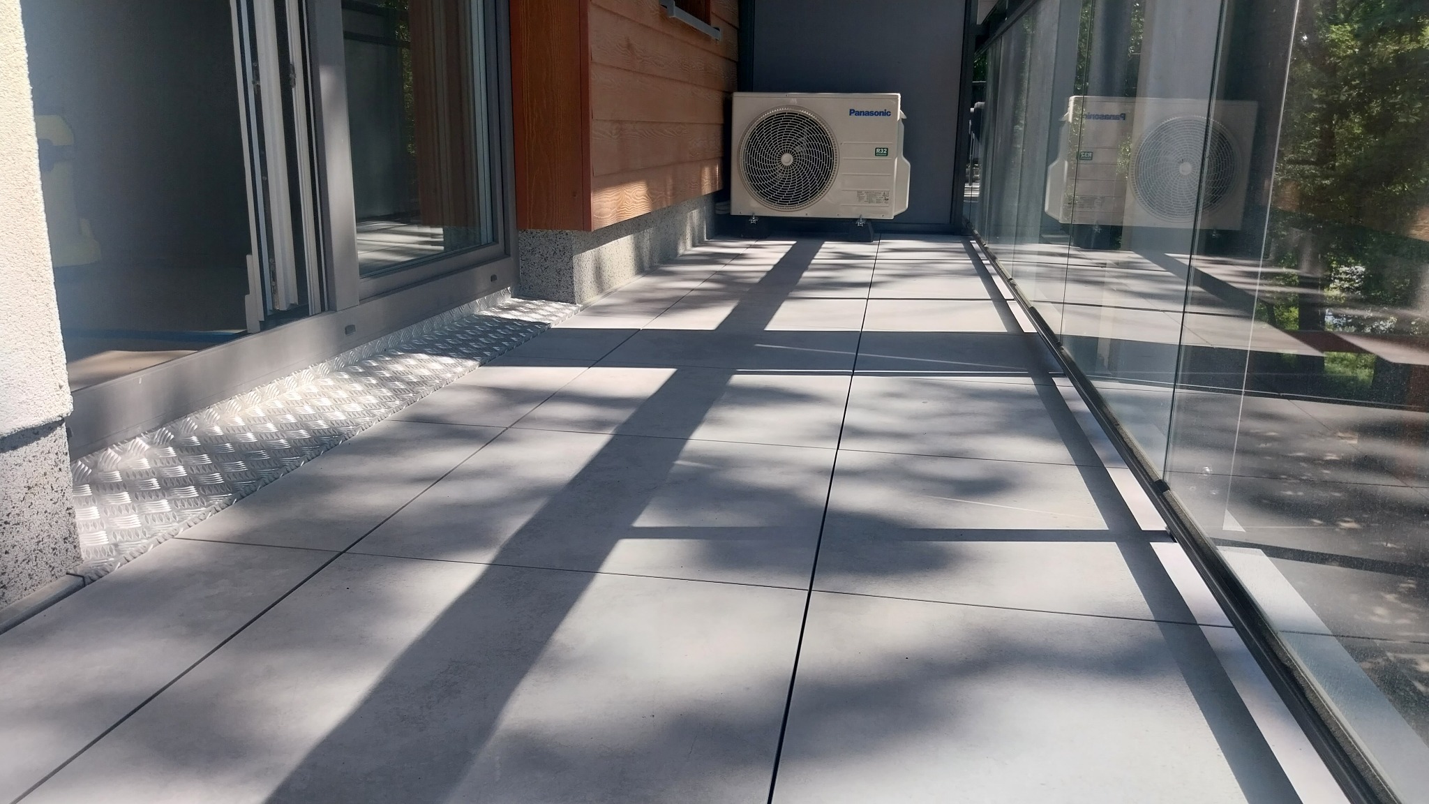 Nowoczesny taras z szarych płyt, z widocznym odpływem liniowym i klimatyzatorem Panasonic. Eleganckie wykończenie z blachy ryflowanej przy drzwiach przesuwnych.