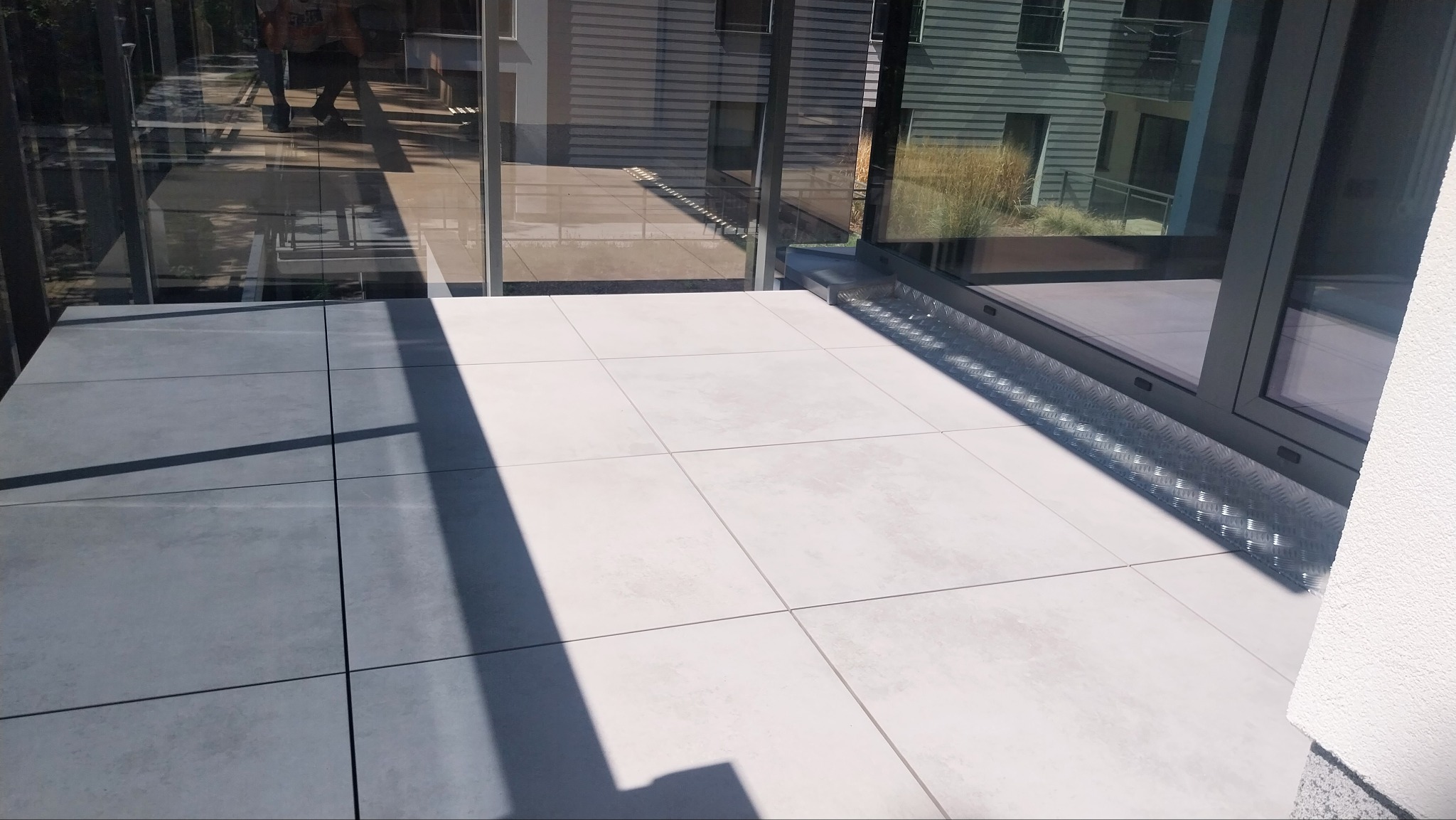 Nowoczesny taras z jasnoszarą, kwadratową płytką, osłonięty szklaną balustradą i dużym oknem. Widoczny fragment elewacji budynku w tle.