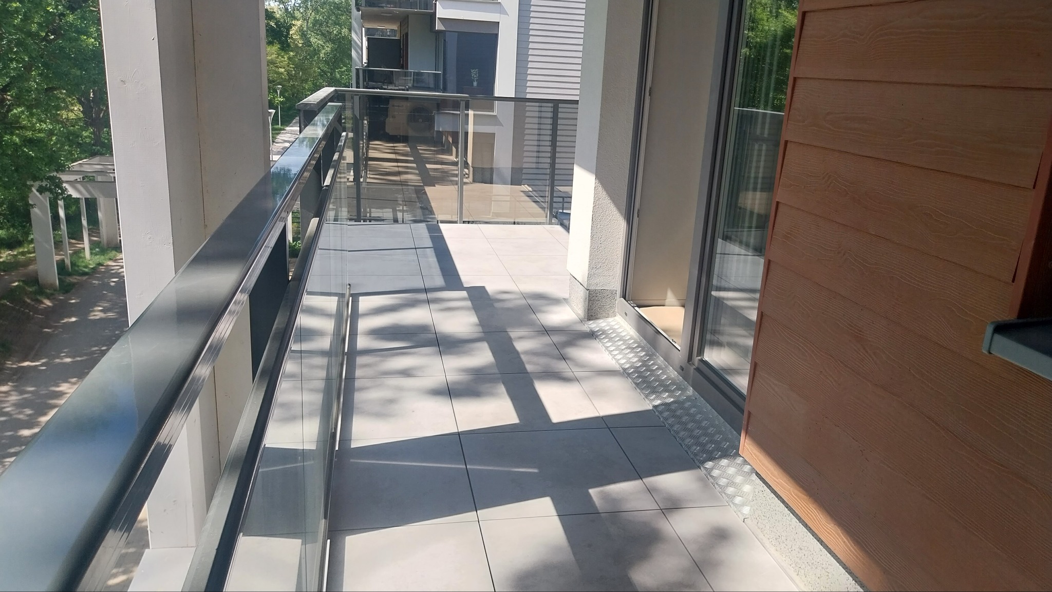 Jasny taras z szarymi płytkami i metalową balustradą z przezroczystym wypełnieniem. Widok na zieleń i architekturę z zewnątrz. Deska elewacyjna z boku.