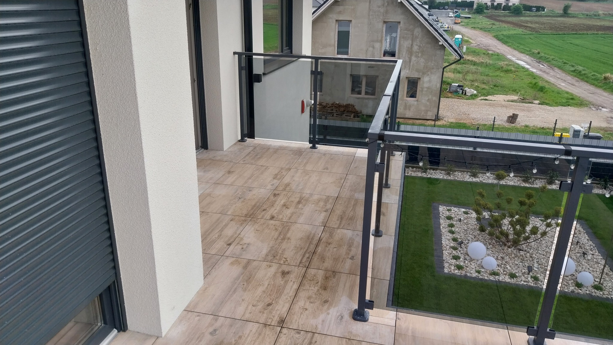 Nowoczesny taras z płytkami imitującymi drewno, szklane balustrady z czarnymi profilami. Widok z góry na starannie zagospodarowany ogród z oświetleniem.