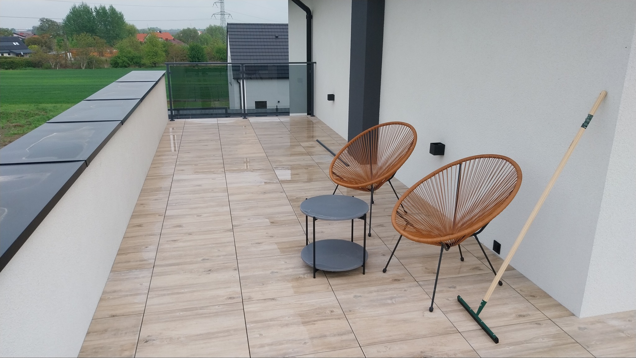 Nowoczesny taras z drewnopodobną płytką, meblami ogrodowymi i szklaną balustradą. Widoczny grabie oparty o ścianę. Taras po deszczu, mokra powierzchnia.