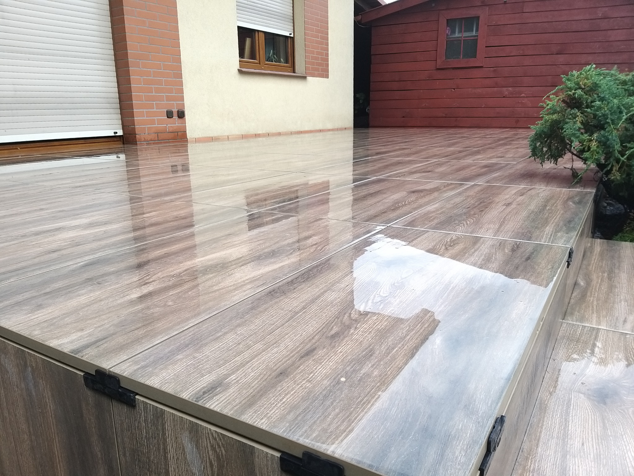 Błyszczący taras z płytek imitujących drewno w Długołęce, z widocznymi detalami łączeń i fakturą. W tle budynek z roletami i drewniany domek.
