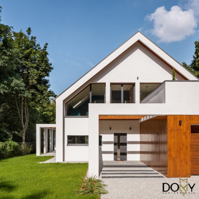 Nowoczesny, biały dom z dwuspadowym dachem, dużymi oknami i drewnianą wstawką garażową, otoczony zielenią i trawnikiem, widok z przodu w słoneczny dzień.