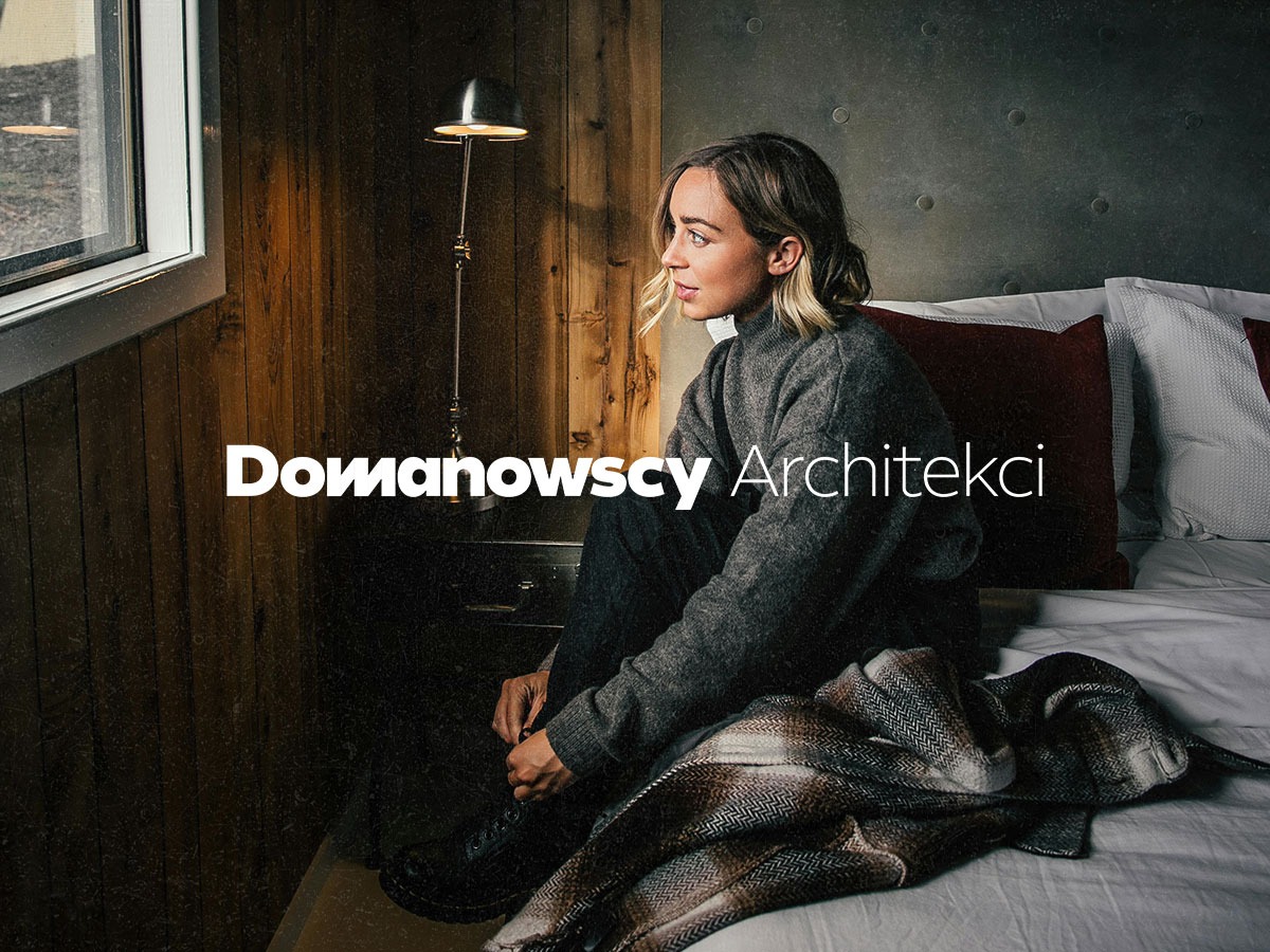 Kobieta siedzi na łóżku w pokoju z drewnianymi ścianami i dużym oknem, spogląda w dal, obok lampa i pled w jodełkę, w tle logo 'Domanowscy Architekci'.