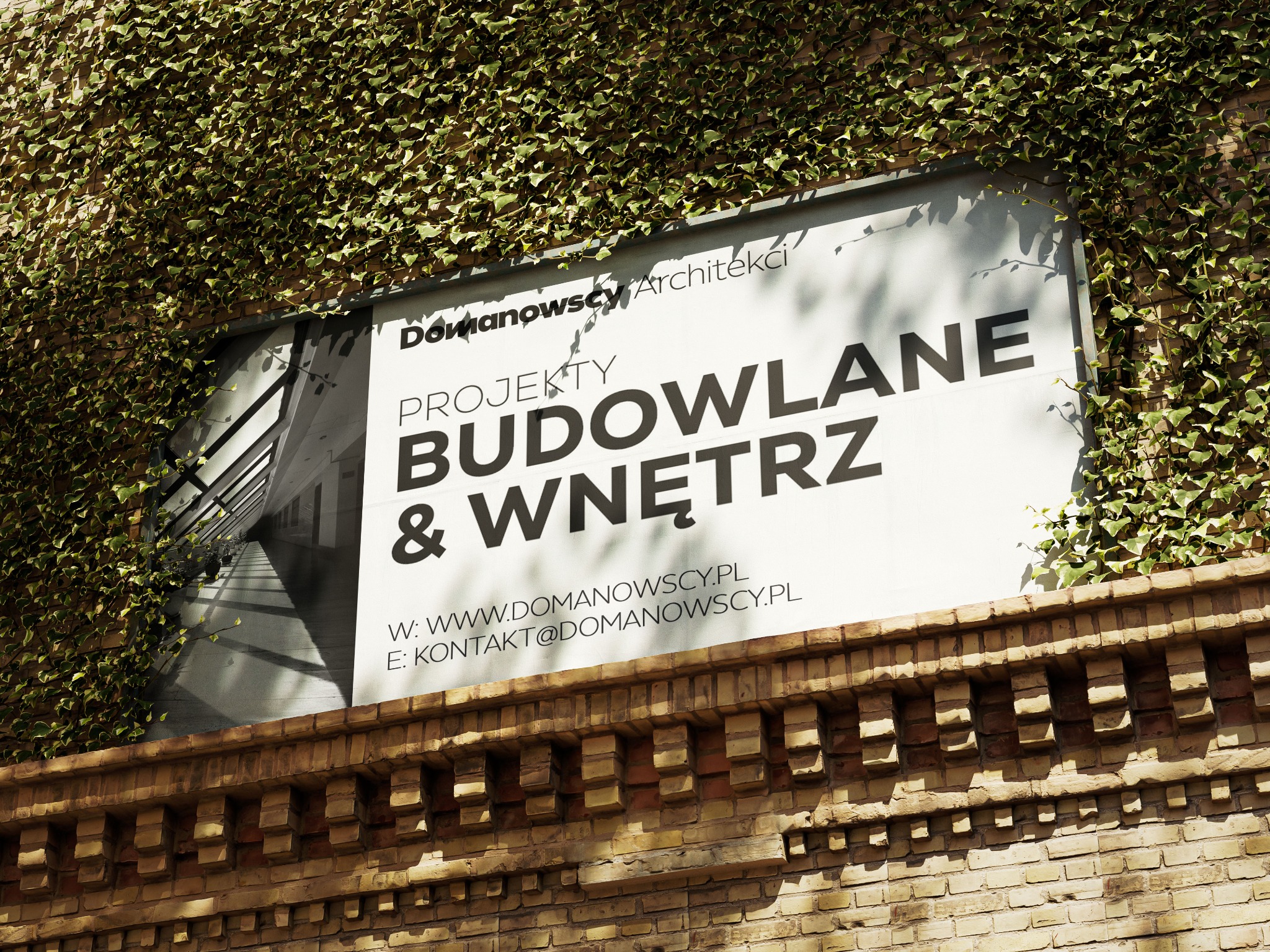 Baner reklamowy firmy architektonicznej 'Domanowscy Architekci' z hasłem 'Projekty Budowlane & Wnętrz' umieszczony na ceglanej ścianie porośniętej bluszczem.