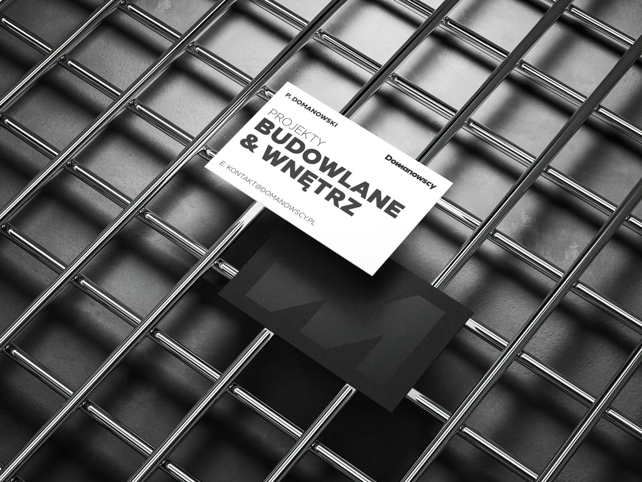 Wizytówki firmy P. Domanowski Projekty Budowlane & Wnętrz na tle metalowej siatki w odcieniach szarości.
