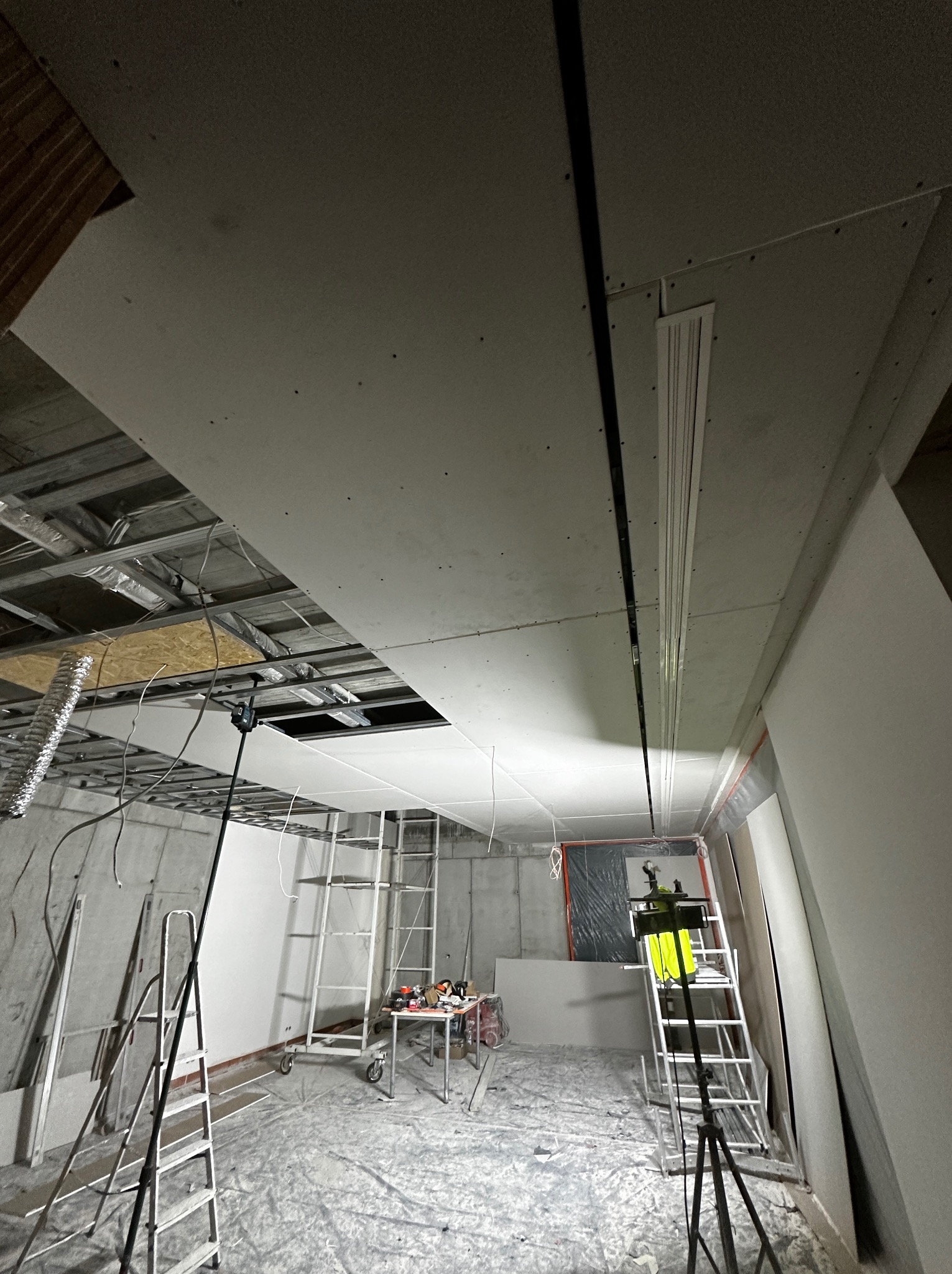 Montaż sufitu podwieszanego z płyt gipsowo-kartonowych w surowym wnętrzu, widoczne rusztowania, narzędzia i instalacje.