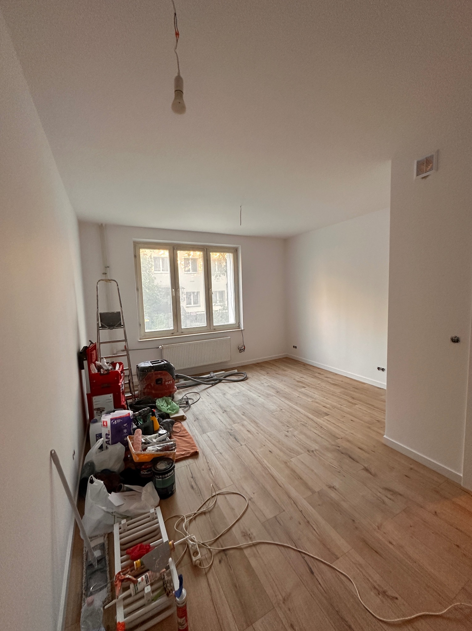 Pokój po odświeżeniu, z nową podłogą i świeżo pomalowanymi ścianami, widoczny sprzęt i narzędzia malarskie, drabina, lampa wisząca na kablu, okno z widokiem na budynek.