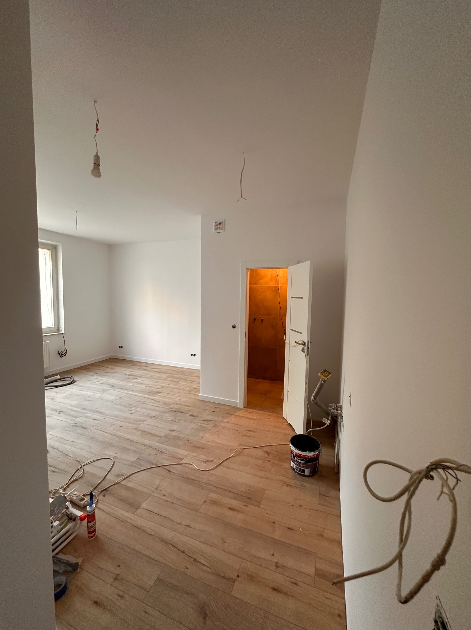 Wnętrze mieszkania w trakcie prac wykończeniowych: świeżo położona podłoga imitująca drewno, pomalowane na biało ściany, otwarte drzwi do łazienki z widocznymi płytkami, wiszące kable elektryczne...