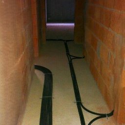 Kamil Sułkowski - Kable elektryczne ułożone w korytarzu w surowym stanie, przymocowane do podłoża metalowymi uchwytami, widoczne ściany z cegły dziurawki.