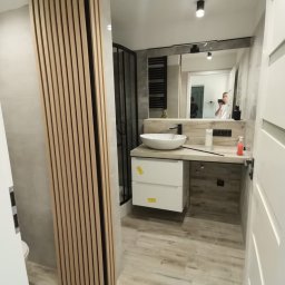 Kamil Sułkowski - Nowoczesna łazienka z drewnianymi lamelami na ścianie, białą umywalką nablatową i kabiną prysznicową z czarnymi profilami, widoczne odbicie osoby robiącej zdjęcie w lustrze.