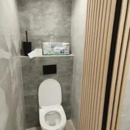 Nowoczesna toaleta po remoncie: szare płytki imitujące kamień, drewniane lamele dekoracyjne, podwieszana muszla klozetowa z czarnym przyciskiem spłukiwania, akcesoria łazienkowe na półce.