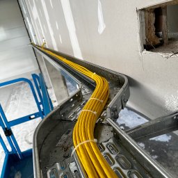 Kamil Sułkowski - Żółte przewody elektryczne ułożone w metalowej korycie kablowej, zamocowanej do ściany w surowym stanie, widoczne elementy rusztowania.