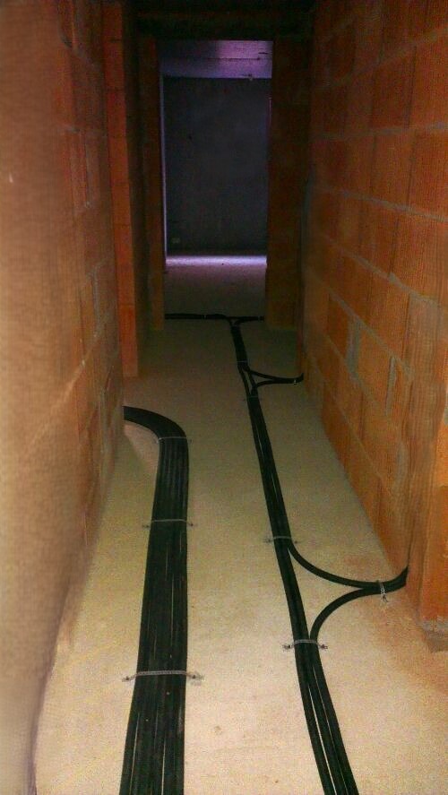 Kable elektryczne ułożone w korytarzu w surowym stanie, przymocowane do podłoża metalowymi uchwytami, widoczne ściany z cegły dziurawki.