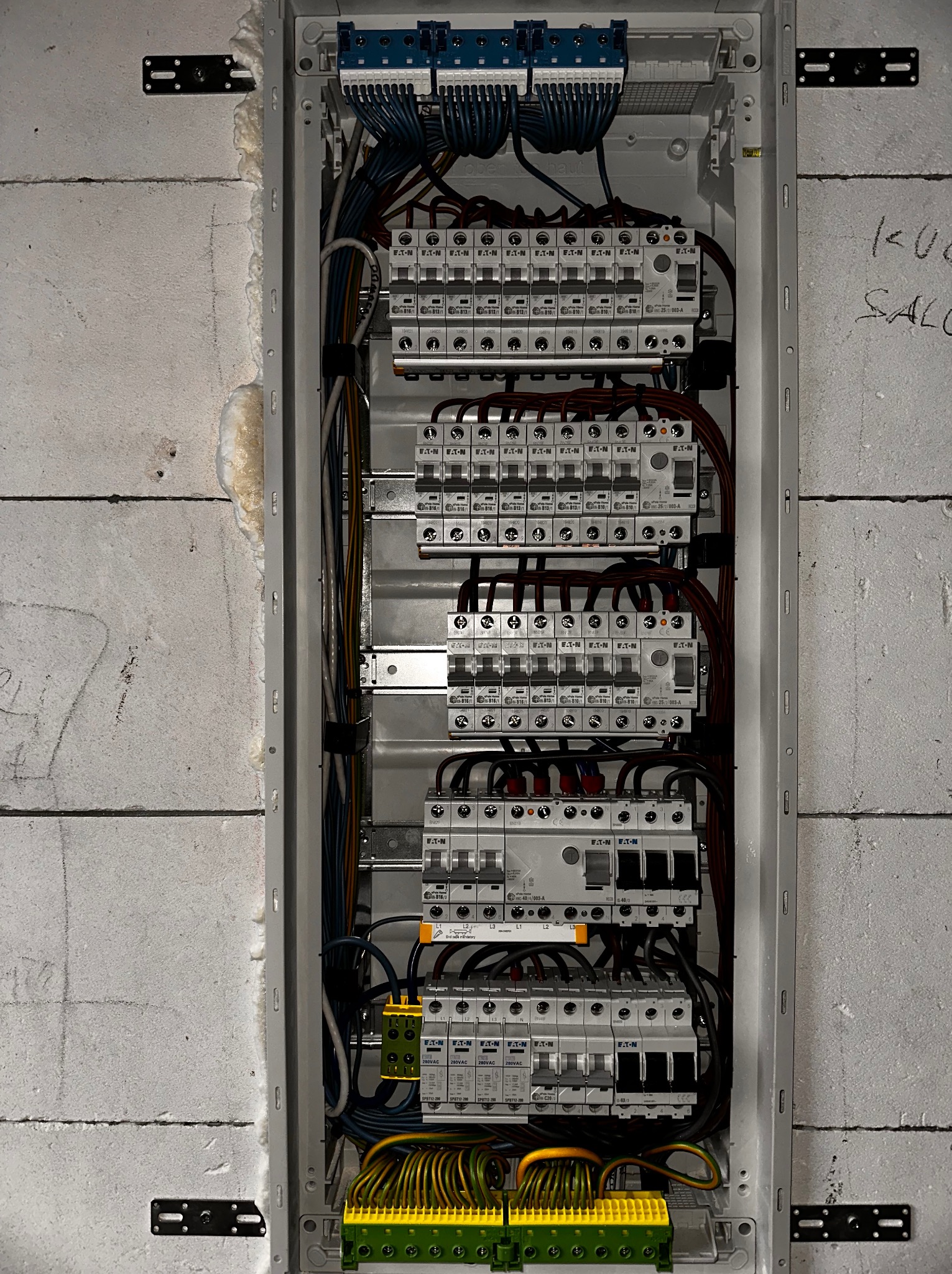Rozdzielnia elektryczna z zainstalowanymi zabezpieczeniami nadprądowymi i przeciwprzepięciowymi marki Eaton, widoczna na tle ściany z betonu komórkowego z widocznymi śladami ołówka.