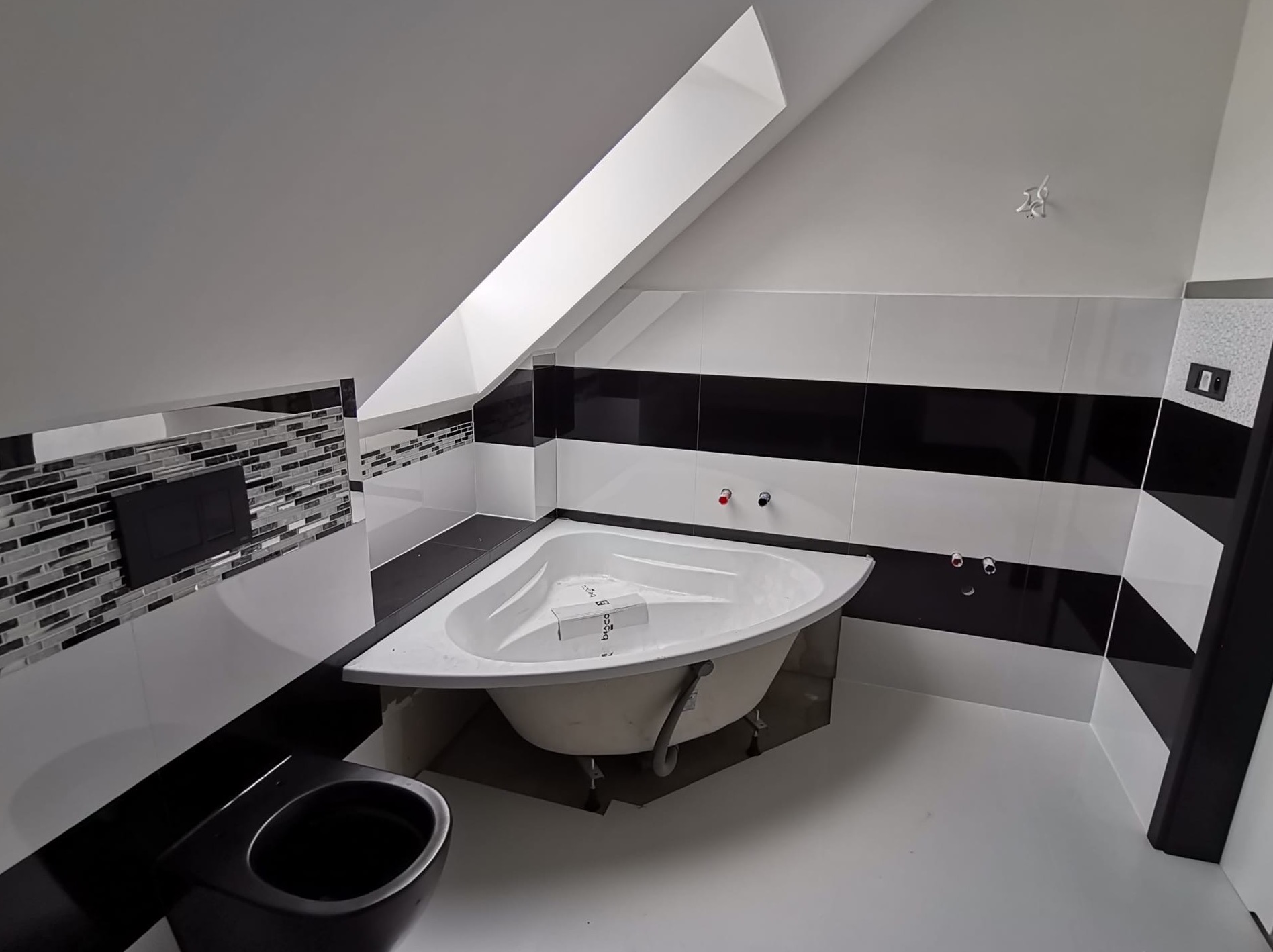 Ukośna łazienka w trakcie wykańczania: narożna wanna z kartonem producenta w środku, czarna miska WC, biało-czarne płytki w pasy i mozaika, surowe przyłącza wodne.