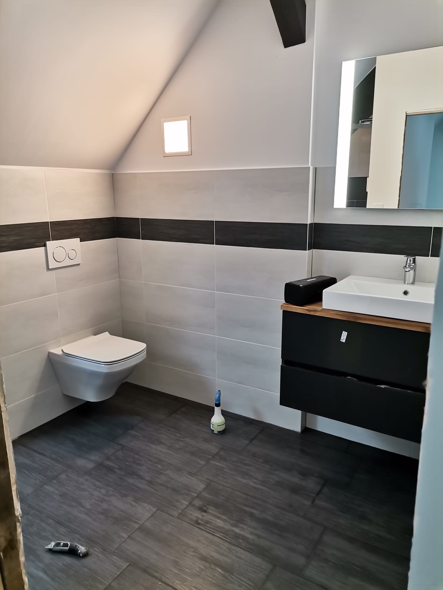 Wykończona łazienka na poddaszu ze ścienną miską WC, umywalką z szafką w kolorze czarnym i szarymi płytkami z czarnym pasem. Na podłodze imitacja drewnianych paneli. Widoczny nóż introligatorski...