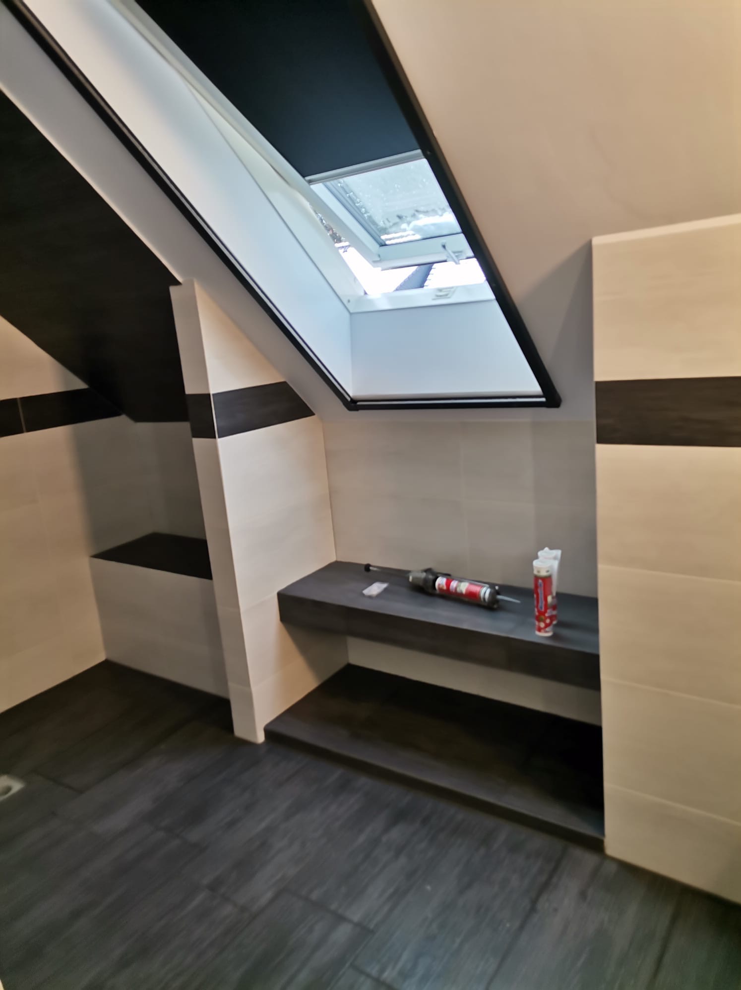 Nowoczesna łazienka na poddaszu z oknem dachowym, wykończona w odcieniach bieli i szarości, z widocznymi narzędziami i tubami silikonu na półce.