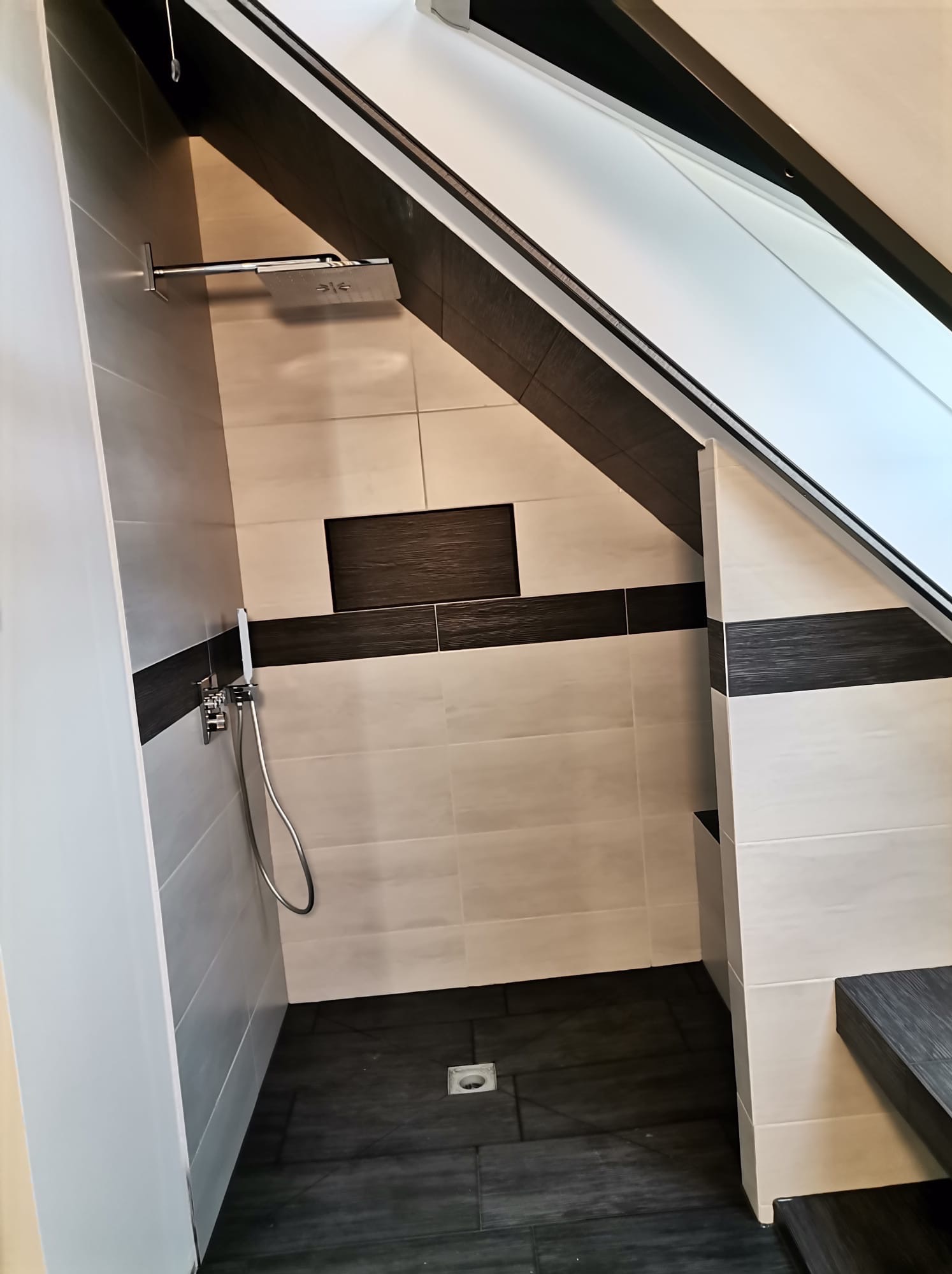 Nowoczesna kabina prysznicowa w łazience na poddaszu, z jasnymi i ciemnymi płytkami, deszczownicą i odpływem liniowym w podłodze.