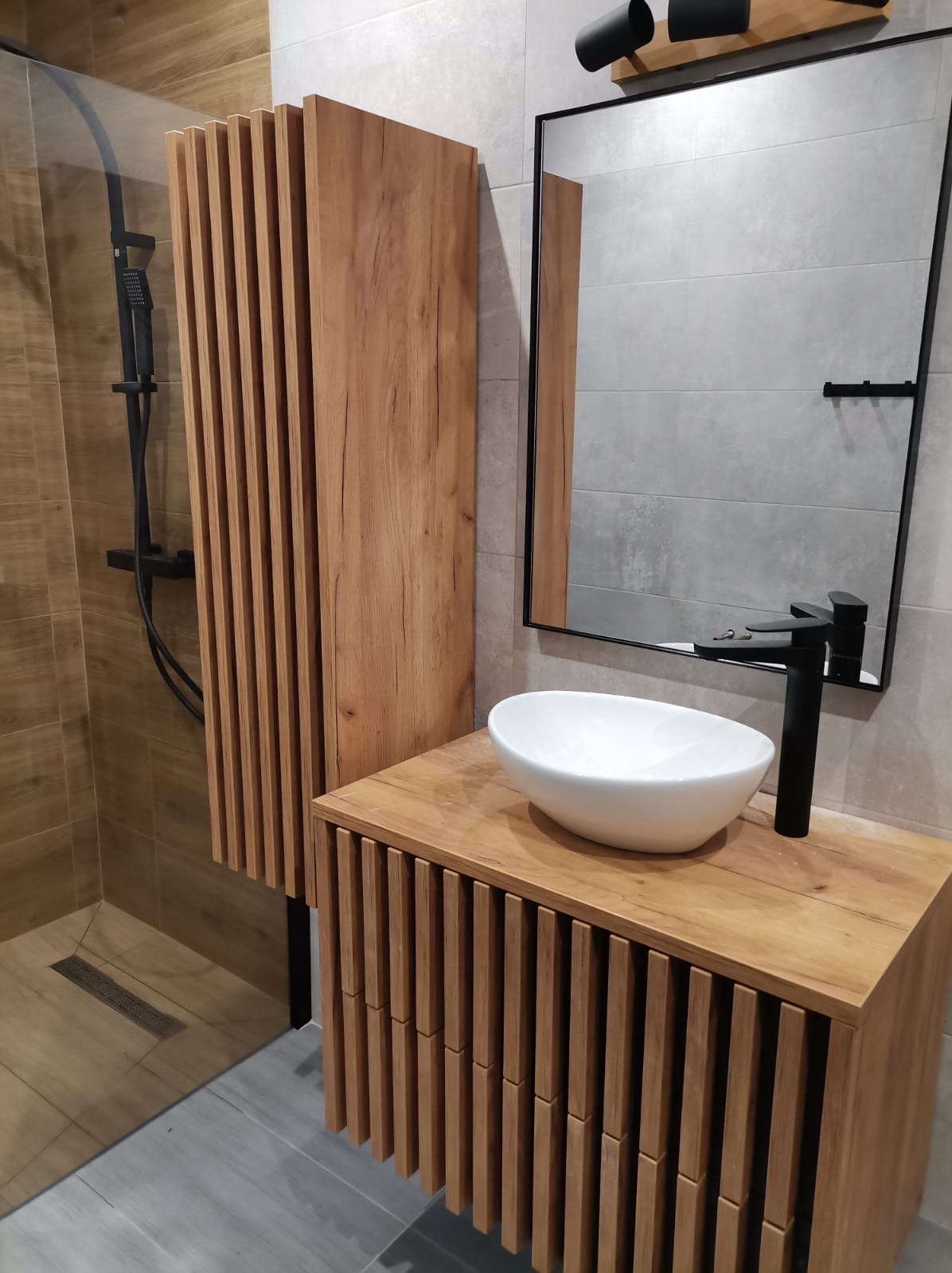 Nowoczesna łazienka z drewnianą szafką pod umywalkę z lamelami, owalną białą umywalką nablatową i czarną armaturą, kabina prysznicowa ze szklaną ścianką.