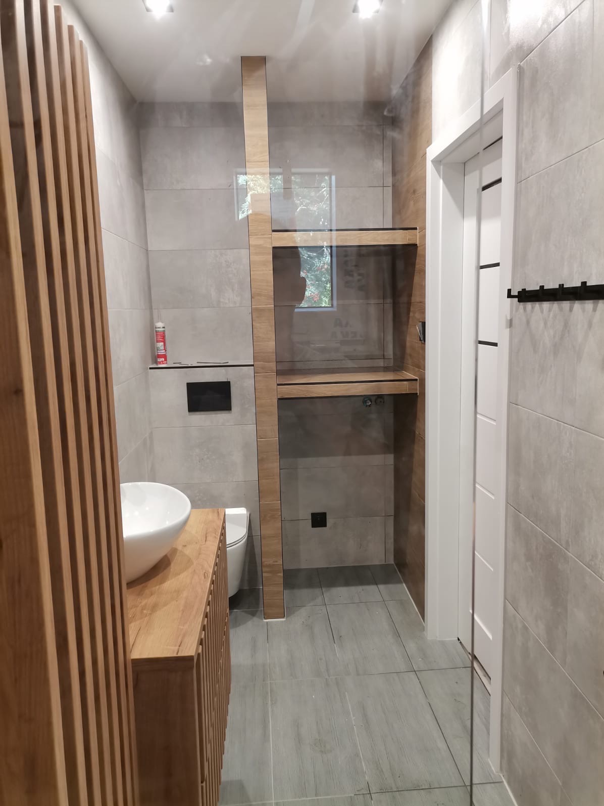 Nowoczesna łazienka z drewnianymi akcentami: umywalka nablatowa, zabudowa z pionowych listew, szklana kabina prysznicowa z drewnianymi półkami, szare płytki na ścianach i podłodze.