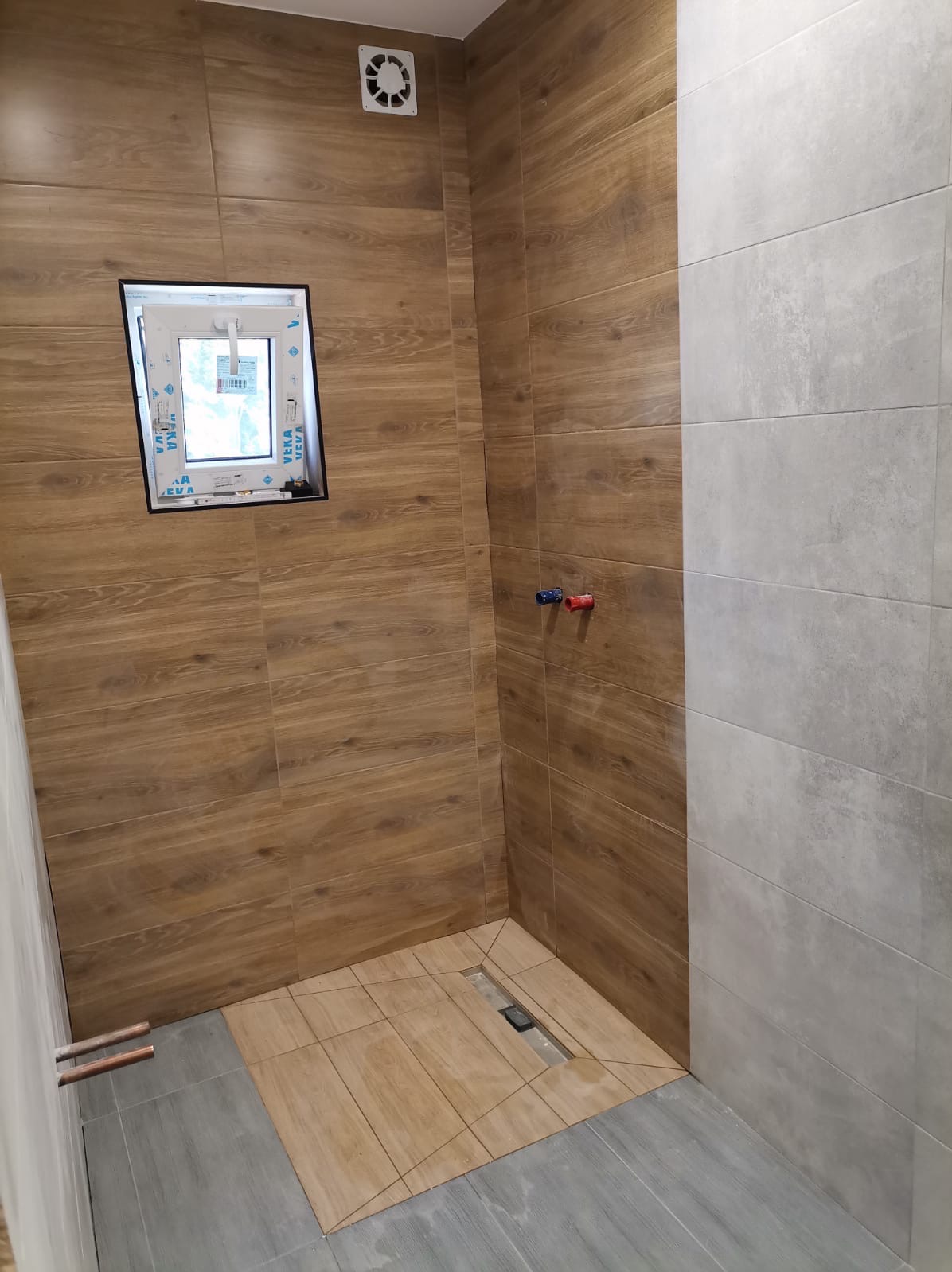Nowoczesna kabina prysznicowa w trakcie wykańczania w Lubsku, z brązowymi i szarymi płytkami imitującymi drewno, widoczny odpływ liniowy i wystające rury.