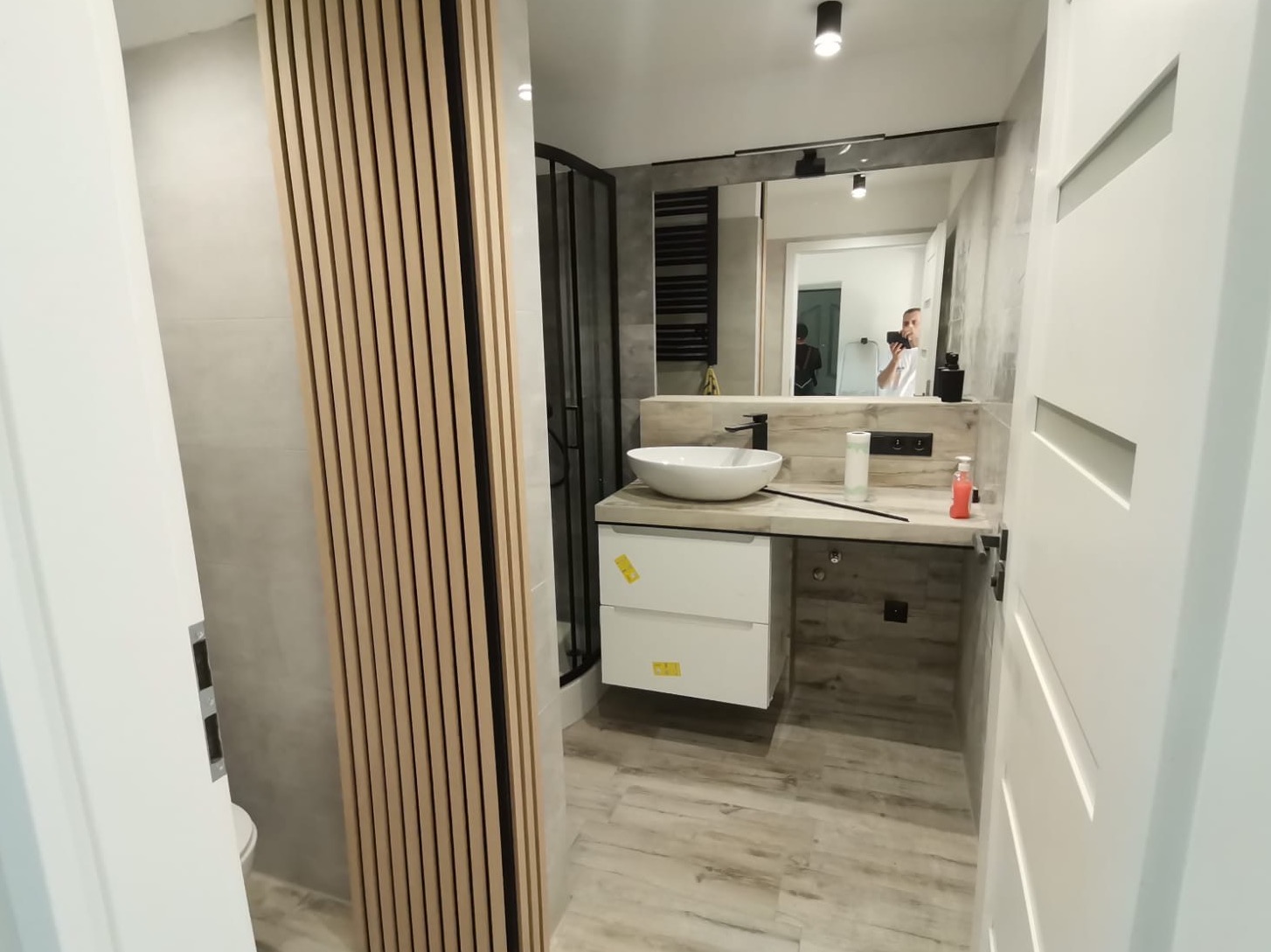 Nowoczesna łazienka z drewnianymi lamelami na ścianie, białą umywalką nablatową i kabiną prysznicową z czarnymi profilami, widoczne odbicie osoby robiącej zdjęcie w lustrze.