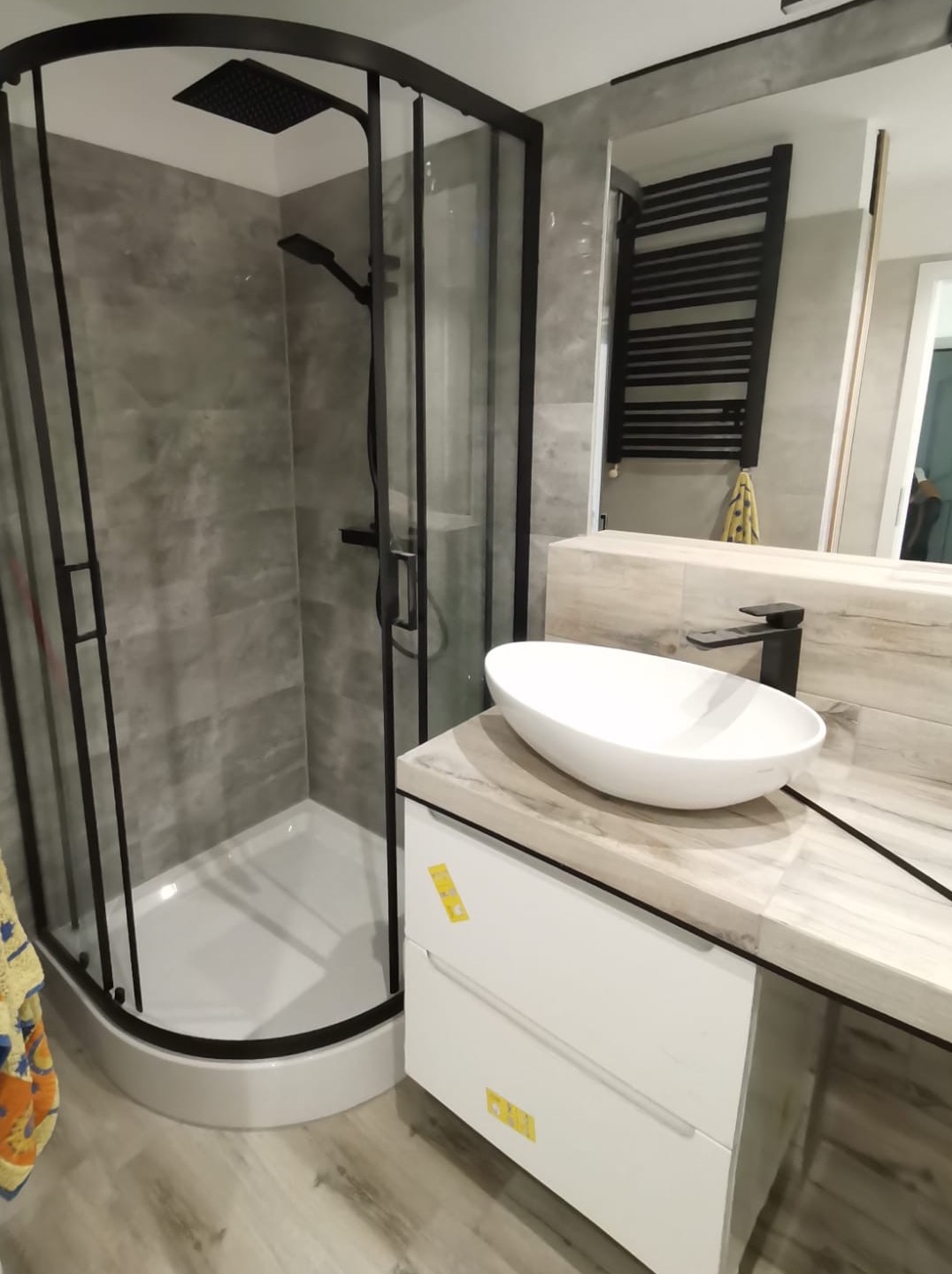 Nowoczesna łazienka z kabiną prysznicową typu walk-in z czarnymi profilami, umywalką nablatową o owalnym kształcie i grzejnikiem drabinkowym w kolorze czarnym.