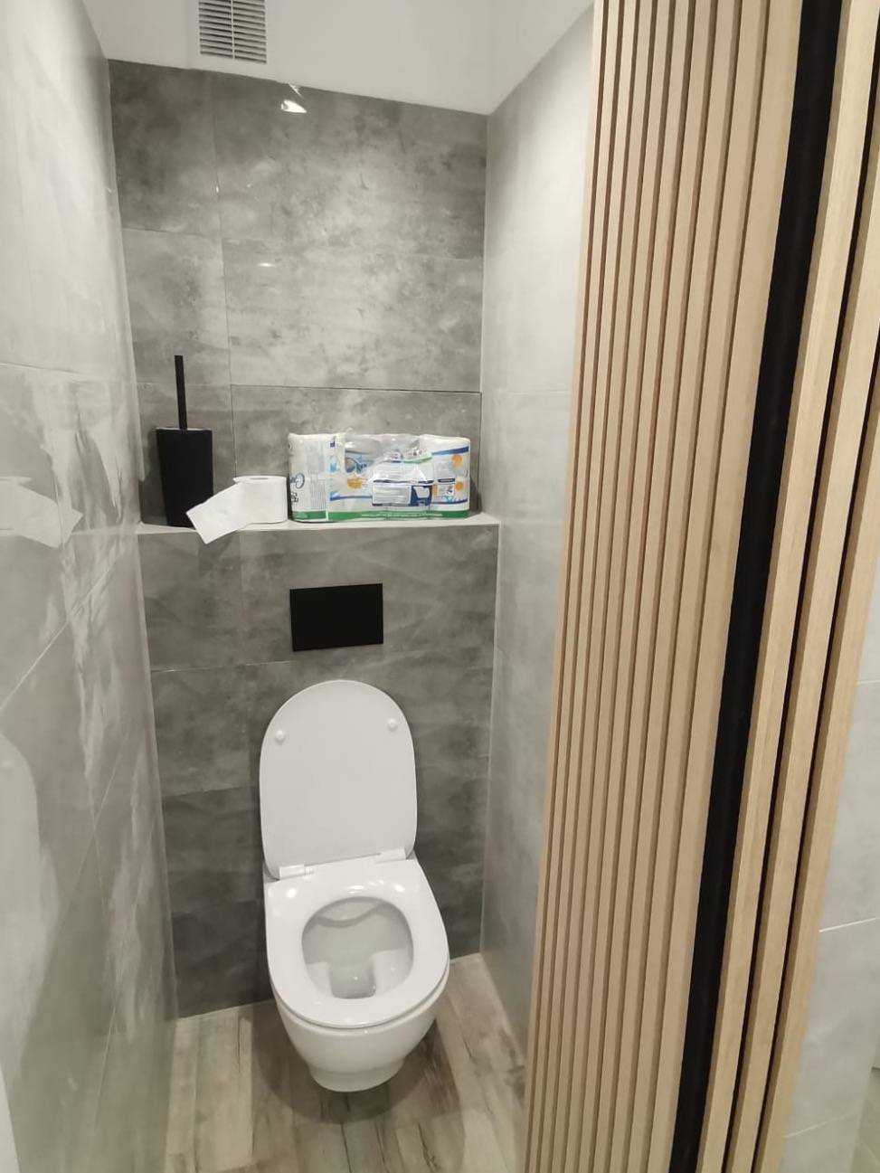 Nowoczesna toaleta po remoncie: szare płytki imitujące kamień, drewniane lamele dekoracyjne, podwieszana muszla klozetowa z czarnym przyciskiem spłukiwania, akcesoria łazienkowe na półce.